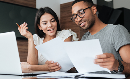 Photo of cheerful loving young couple using laptop and analyzing their finances with documents. Look at papers.