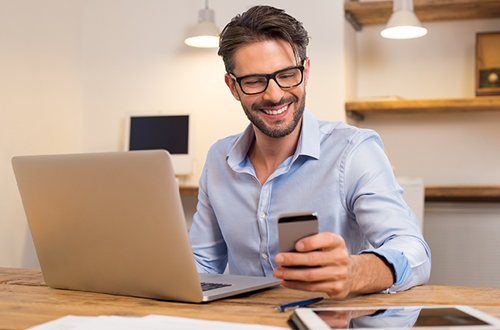 Young happy businessman smiling while reading his smartphone. Portrait of smiling business man reading message with smartphone in office. Man working at his desk at office.