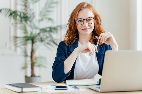 Positive red haired young female entrepreneur watches business webinar, writes email, makes notes in notepad, wears optical glasses, dressed in formal clothes, works online, involved in banking sphere